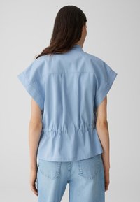 Lichtblauwe blouse met korte, brede mouwen, een gathered taille en een gladde textuur. Gecombineerd met lichte denim jeans, met achteraanzicht.