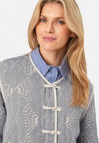 Blau gemusterte Jacke mit beiger Stickerei, darunter ein hellblaues kragenloses Hemd. Auffällige Kordelverschluss an der Jacke.