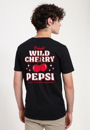 Ung mand med krøllet hår iført en sort T-shirt med et rødt kirsebærmotiv og teksten "Drink! Wild Cherry Pepsi" på bagsiden.