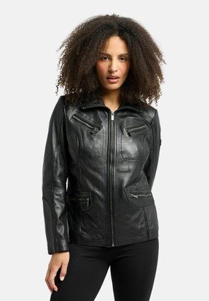 Vrouw met krullend haar die een zwarte leren jas draagt met ritszakken en een zwarte broek op een effen lichte achtergrond.