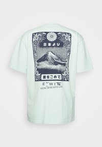 T-shirt vert clair à manches courtes avec un graphisme bleu marine du Mont Fuji, une bordure florale, du texte japonais, et l'inscription "DEPUIS LE JAPON AVEC AMOUR" au dos.