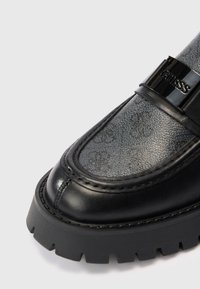 Musta loaferit, joissa on teksturoitu pintamateriaali ja kiiltävä musta nauha sekä rohkeat brändäykset. Paksu kumipohja, jossa on uratehoste. Klassinen muotoilu.
