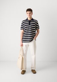 Marineblå og kremstripet polo-skjorte med krage og lomme. Kombinert med hvite bukser, brune sko og holder en beige totebag.