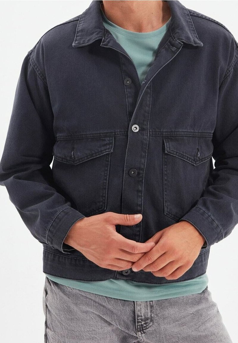 Dunkle Jeansjacke mit klassischem Kragen, zwei Brusttaschen, Knöpfen vorne und langen Ärmeln. Darunter ein hellblaues T-Shirt.