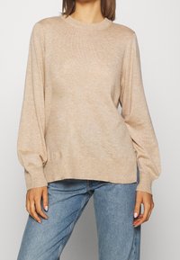 Pull en tricot beige avec col rond côtelé et manches bouffantes. Texture douce, coupe décontractée, associé à un jean en denim bleu clair.