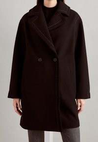 Cappotto nero doppiopetto con ampi rever, cuciture bianche in evidenza, tasche laterali e vestibilità rilassata. Tessuto di lana morbido.