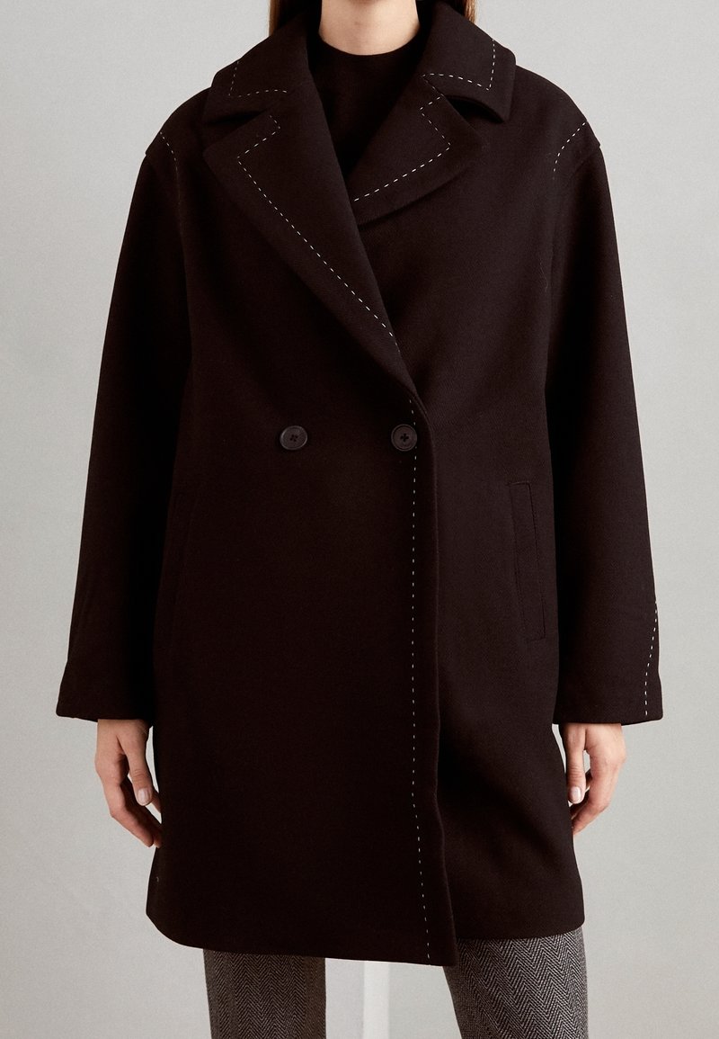 Cappotto nero doppiopetto con ampi rever, cuciture bianche in evidenza, tasche laterali e vestibilità rilassata. Tessuto di lana morbido.