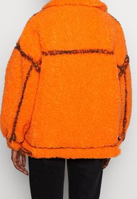 Oranžová fleecová bunda s texturovaným povrchem, s kontrastním černým prošíváním a límcem. Volný a nadrozměrný střih.