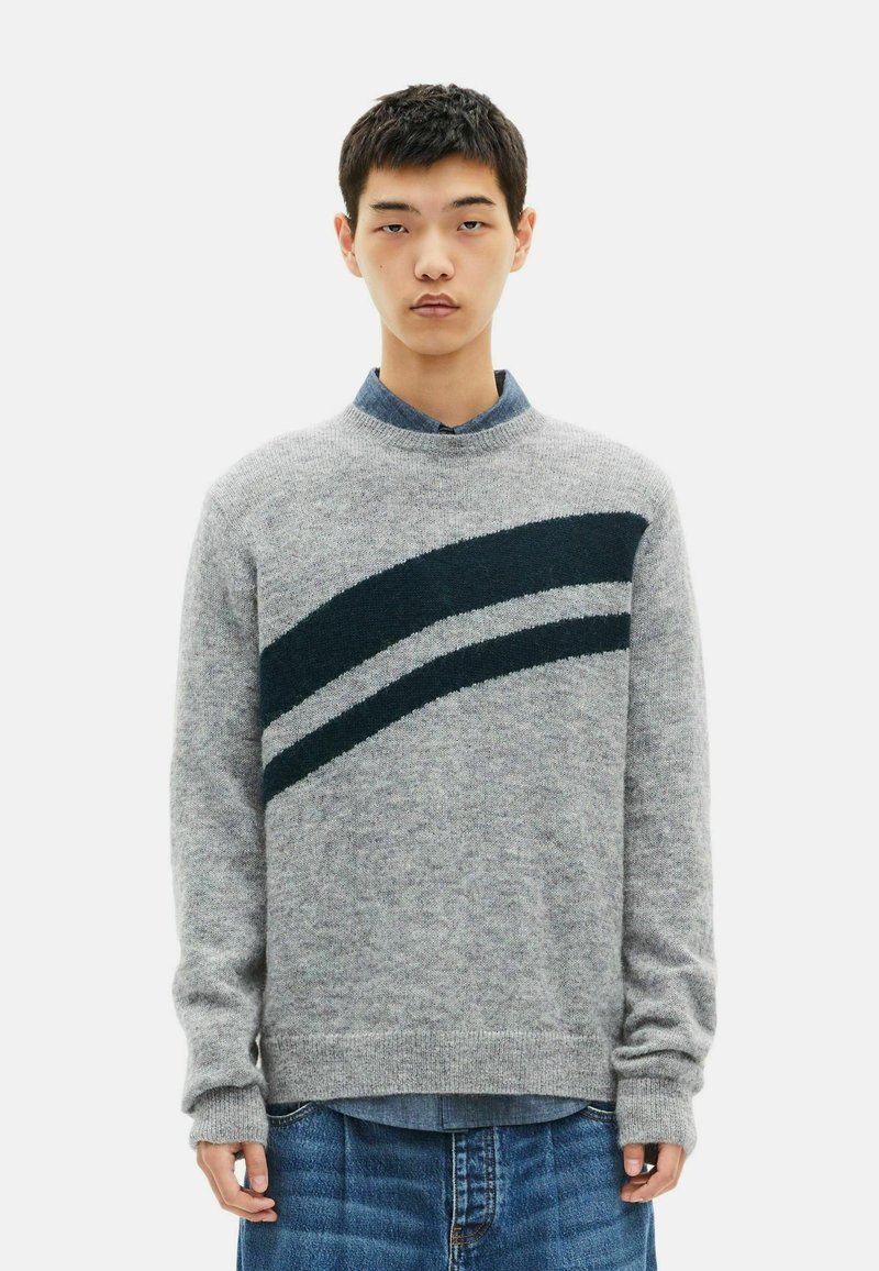 Junger Mann, der einen grauen Pullover mit zwei dunklen diagonalen Streifen über einem blauen Hemd mit Kragen und blauen Jeans trägt, steht vor einem weißen Hintergrund.
