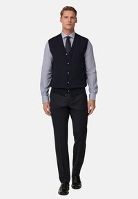 Námořnická vlna vesta přes pruhovanou modrou košili, kombinovaná s tmavými kalhotami a černými botami. Zapínání v přední části a čisté linie definují tento outfit.