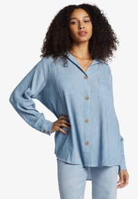 Light blue striped button-up shirt with a collar, two front pockets, and long sleeves, featuring wooden buttons and a relaxed fit.