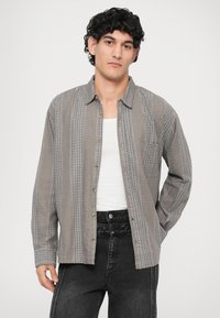 Camicia a quadri grigia con design a bottoni, taschino singolo sul petto e tessuto testurizzato; abbinata a jeans neri.
