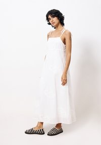 Weißes Baumwoll-Maxi-Kleid mit verstellbaren Trägern und floralen Stickereien. Kombiniert mit schwarz-beigen gestreiften Slippern.