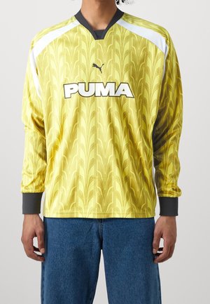 Osoba ubrana w żółtą sportową koszulkę z długim rękawem marki Puma z wzorzystym wzorem i białymi akcentami na ramionach, w połączeniu z niebieskimi dżinsami.