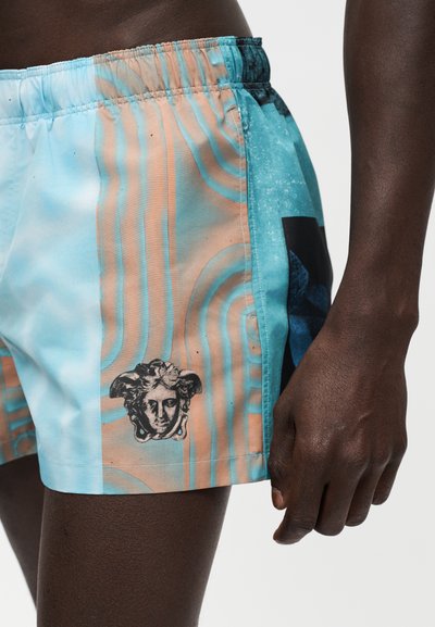 Personne portant un short de bain géométrique bleu pastel et orange avec un motif de tête de style Méduse sur la jambe gauche, debout avec le bras le long du corps.