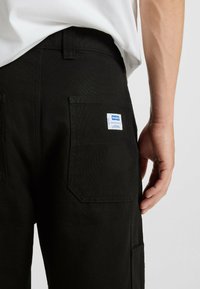 Close-up of a person wearing black pants with a back pocket displaying a white and blue "Hugo" label, and a white shirt tucked in.