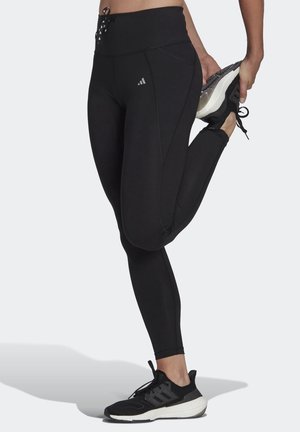 Leggings sportivi neri con vita alta, caratterizzati da dettagli cuciti e un logo, abbinati a scarpe sportive nere e texture visibili.