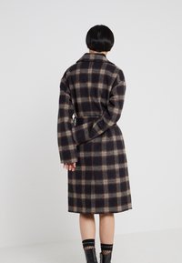 Abrigo largo de cuadros en tonos negro, rojo y beige, con cintura ceñida con cinturón, hombros caídos y mangas anchas. La tela parece de lana y con textura.