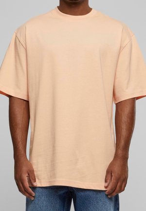 Homme portant un t-shirt ample à manches courtes couleur pêche clair et un jean bleu sur un fond gris uni.