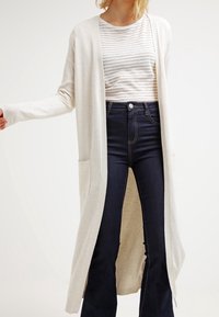 Beige lång kofta med öppen framdel och två sidofickor, bärs över en randig topp och mörka, högmidjade utsvängda jeans. Mjuk textur.