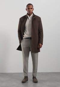 Hnědý kabát s vysokým límcem a předním zipem, nošený přes světle šedý svetr a šedé kalhoty. Hnědé semišové boty s minimalistickým designem.
