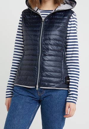 Person iført en navyblå quiltet, ærmeløs hættetrøje over en langærmet hvid og navystribet skjorte med blå jeans.