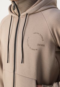 Sweat à capuche beige zippé avec cordons noirs, texte circulaire « ARCTIC XTREME » et logo « BOSS » sur la poitrine, porté par-dessus un pull à col roulé côtelé.