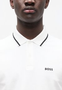 Λευκό πόλο με πλεκτό γιακά που έχει μαύρες ρίγες. Περιλαμβάνει μικρό μαύρο κεντητό λογότυπο "BOSS" στην αριστερή πλευρά.