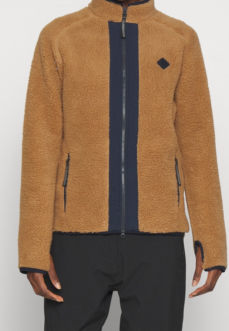 Brązowa polarowa kurtka z niebieską pionową paskiem, wysoki kołnierz, zamek błyskawiczny oraz zamykane na zamek kieszenie boczne. Posiada logo w kształcie diamentu na klatce piersiowej.