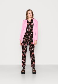 Blazer rosa su una tuta nera con stampa floreale, caratterizzata da fiori rosa e bianchi. L'outfit è abbinato a sandali platform neri e spessi.