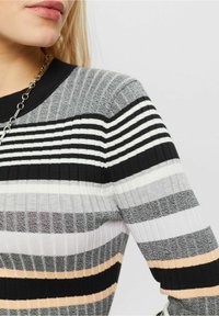 Pull en maille côtelée à rayures grises, noires et blanches avec des accents pêche. Présente un col rond et un tissu texturé. Collier chaîne visible.