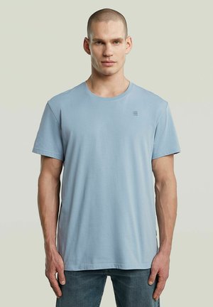 T-shirt en coton bleu clair avec des manches courtes et un col rond. Présente un petit logo imprimé sur la poitrine. Texture lisse et coupe décontractée.