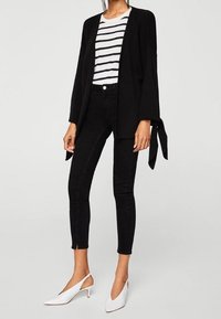 Cardigan noir à manches longues et ouvert avec détails noués, T-shirt rayé blanc et marine, jean skinny noir, et escarpins blancs à bride arrière.