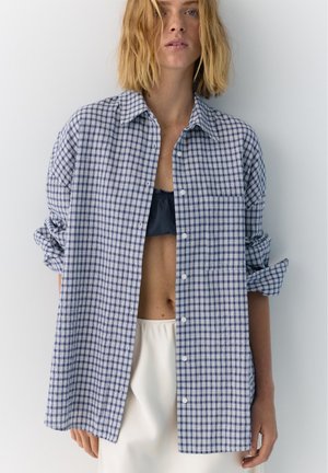 Femme aux cheveux blonds mi-longs portant une chemise ample à carreaux bleus et blancs par-dessus un crop top noir et un pantalon blanc, debout contre un mur blanc.