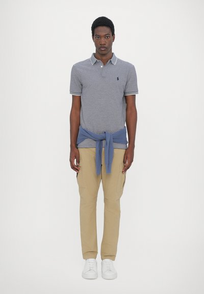Camisa polo de rayas azul marino con cuello, suéter azul atado a la cintura, pantalones cargo beige y zapatillas blancas.