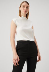 Hvit ermeløs bluse med høy krage, glatt tekstur, kombinert med svarte skreddersydde bukser; modellen har på seg minimalistiske øredobber.