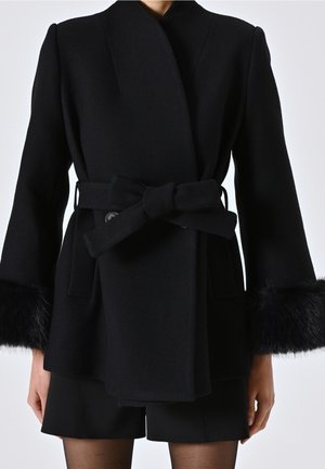 Black belted wool coat with fur cuffs worn over black shorts and sheer tights, shown from neck to mid-thigh on a person.