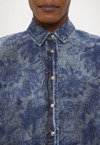 Denimskjorte med et blått blomstermønster og gullknapper foran, strukturert krage og myk tekstur.
