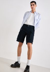 Shorts cargo bleu marine avec plusieurs poches, associés à une chemise rayée bleu clair et un débardeur blanc. Des chaussures noires et des chaussettes blanches complètent le look.