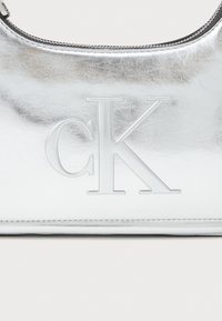 Geantă metalică argintie cu logo-ul „CK” embosat, textură netedă, formă rotunjită și închidere cu fermoar pe marginea superioară.