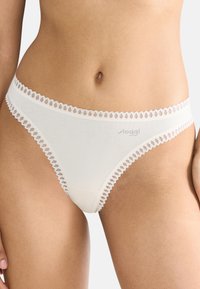 Kvinna klädd i vitt bikiniunderklädesset med spetskant och "sloggi"-logotyp på höften, visad från midjan till övre låren.