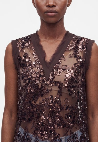 Gros plan d'une personne portant un haut sans manches transparent avec des motifs floraux en sequins marron et un col en V aux bords effilochés.