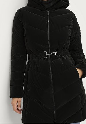 Manteau matelassé noir avec capuche, ceinture élastique avec boucle argentée, fermeture éclair à l'avant et une main dans la poche, porté sur un haut noir.