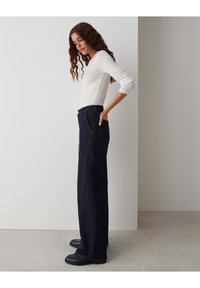 Dark blue, wide-leg jeans with contrasting orange stitching, high waist, and side pockets. Paired with a long-sleeve white top and black shoes.