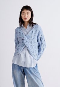 Gaiši zils adījuma džemperis ar brīvu piegriezumu, V-veida kakla izgriezumu un sarežģītu rakstu. Valkāts virs gaiši zila krekliņa un kombinēts ar džinsu biksēm.