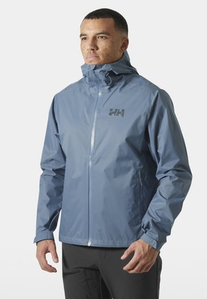 Man met kort haar en sproeten, gekleed in een lichtblauwe waterdichte jas met capuchon en ritszakken, staand tegen een witte achtergrond.