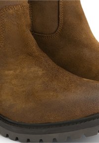 Gros plan de bottines en daim marron avec des semelles épaisses en caoutchouc noir et des panneaux latéraux élastiques.