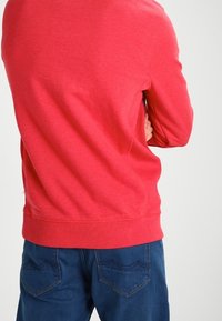 Sudadera de algodón roja con cuello redondo, puños y bajo acanalados. Combinada con vaqueros azules que tienen bolsillos traseros y detalles sutiles de costura.