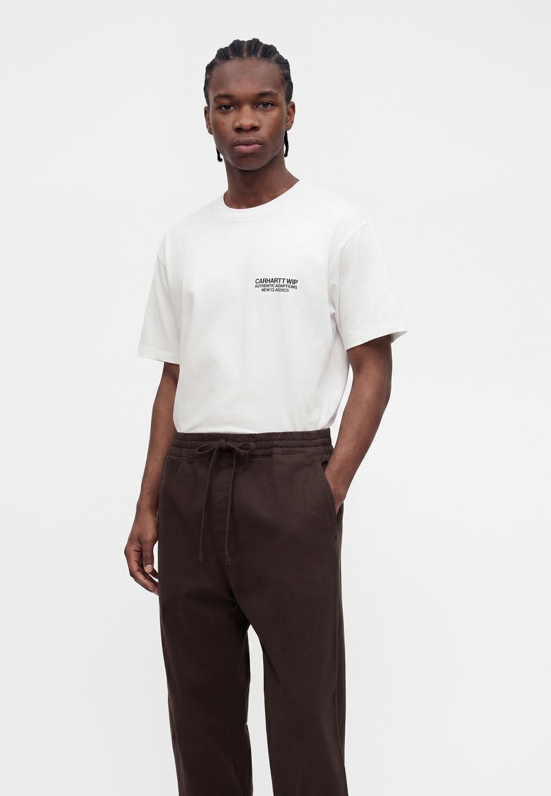 Giovane uomo con capelli intrecciati indossa una t-shirt bianca Carhartt WIP e pantaloni marrone scuro con coulisse, in piedi davanti a uno sfondo grigio uniforme.