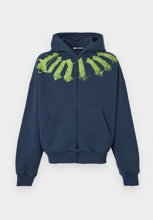 Mand med krøllet hår og solbriller iført en navy hættetrøje med stor grøn tekst, beige vidde bukser og sort-hvide sneakers.